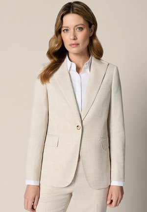 Kobieta o jasnobrązowych włosach, ubrana w beżową marynarkę i dopasowane spodnie, pod którą ma białą koszulę z kołnierzykiem, stoi na tle jednolitego tła.