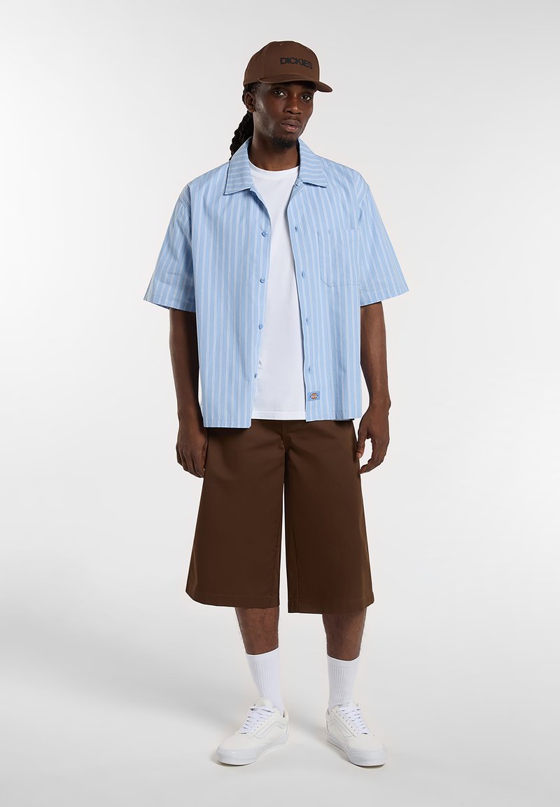 Blå og hvide stribede kortærmede skjorte, hvid T-shirt under, brune knælange shorts, hvide sneakers og en brun kasket med logo.