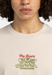 Béžové bavlnené tričko s grafikou troch lebiek v klobúkoch v zelenej a červenej farbe a textom „The Doers“ a „BUILD SOMETHING“ pod nimi.