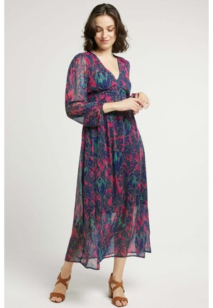 Robe maxi en tissu transparent avec un motif floral dans les tons de violet, rose et vert. Encolure en V, manches longues et silhouette fluide. Sandales à talons bruns.