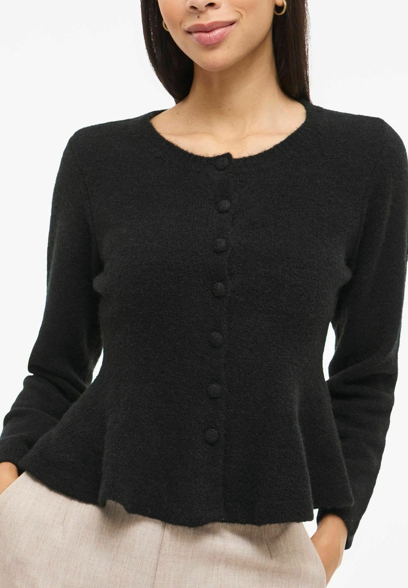 Melns kardigans no mīksta, strukturēta auduma, ar apaļu kakla izgriezumu, priekšā aizpogājams un ar uzplečiem paplašinātu apakšu.