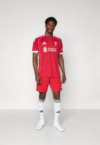 Echipament de fotbal roșu Liverpool cu accente albe, având guler rotund, logo și print al sponsorului; asociat cu pantaloni scurți roșii și șosete albe cu dungi negre.