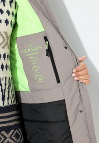 Giacca grigia e nera con una fodera trapuntata verde; presenta una tasca con zip etichettata con icone meteorologiche per impermeabile, traspirante e antivento.