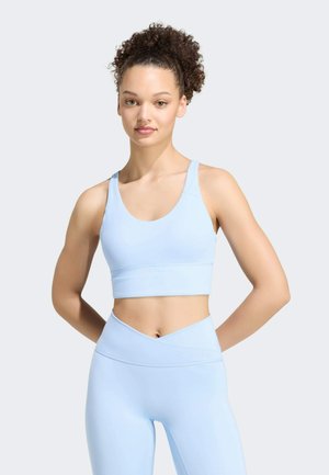 Giovane donna con capelli ricci che indossa un reggiseno sportivo azzurro chiaro e leggings a vita alta, in piedi con le mani dietro la schiena su sfondo semplice.