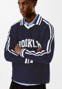 Námořnický pletený svetr s bílými pruhy, výstřihem do V a nápisem "BROOKLYN" na hrudi. Žebrovaná textura a volný střih.