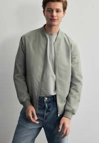 Lichtgrijze bomberjack, zachte textuur, ritssluiting, geribde manchetten, over een grijze t-shirt met blauwe jeans met een hoge taille en knoopdetails.