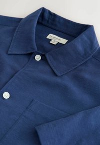 Tumesinise lühikeste varrukatega särk, millel on nööbitav krae, tekstureeritud kangas ja üks rinnatasku. Varustatud valgete nööpidega.