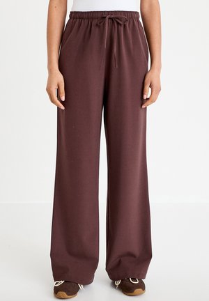 Osoba ubrana w ciemnobrązowe spodnie o szerokich nogawkach z paskiem ściągającym w pasie oraz brązowe buty sportowe, stojąca na jasnym tle.