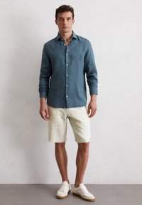 Blaues Leinenhemd mit langen Ärmeln und Knopfverschluss, kombiniert mit weißen Leinen Shorts und cremefarbenen Sneakers. Einfaches Design, lockere Passform.