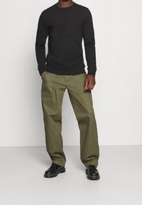 Svart långärmad tröja med ribbade ärmslut, kombinerad med olivgröna byxor med framfickor. Svarta skor med rund tådesign.