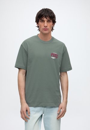 Giovane uomo con capelli scuri che indossa una maglietta verde larga con piccolo testo rosso e bianco sul petto e jeans azzurri.