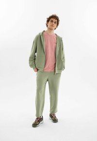 Ljusgrön zip-up hoodie med fickor, kombinerad med matchande ljusgröna sweatpants. Bärs över en rosa skjorta. Sneakers med svarta och orangea accenter.
