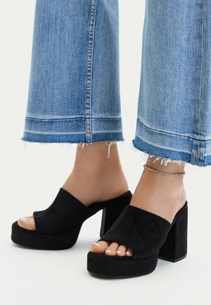 Füße in schwarzen Wildleder-Plattform-Peeptoe-Pumps mit klobigen Absätzen, kombiniert mit blauen Jeans mit ausgefranstem Saum und einem dünnen Knöchelarmband am rechten Fuß.