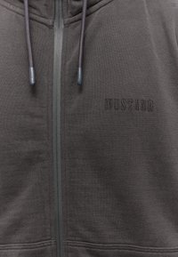 Cárdigan con cremallera de color gris oscuro, hecho de una tela suave, que presenta una cremallera en contraste, detalles de cordón y el logo en relieve "MUSTANG" en el pecho.