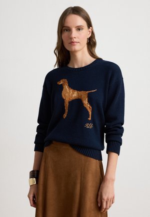 Donna che indossa un maglione blu navy con la silhouette di un cane marrone e una gonna marrone, in piedi contro uno sfondo chiaro uniforme.