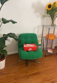 Zelené kordurové lounge kreslo s prešívaným dizajnom, chrome hviezdicovým základom a červenou krabičkou na topánky Nike na sedadle, na pozadí svetlej steny.