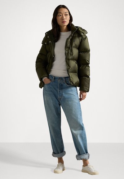Donkergroene pufferjack met een rits en drukknoopsluiting, voorzien van een capuchon en een gladde textuur, gecombineerd met lichtblauwe ruime jeans en witte sneakers.