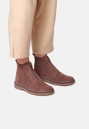 Bottines en daim marron avec fermetures éclair à l'avant, portées avec un pantalon beige raccourci sur un fond clair.