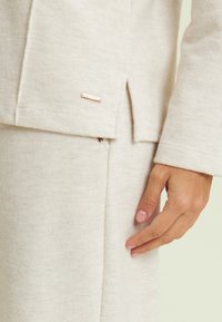 Maglione di maglia beige chiaro con texture a coste, caratterizzato da uno spacco laterale e un piccolo accento in metallo vicino all'orlo.