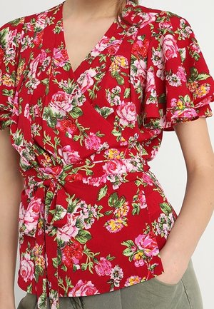 Blouse portefeuille rouge à imprimé floral rose et jaune, manches volantées et ceinture nouée à la taille, portée avec un pantalon vert.