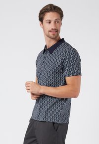 Kortärmad pikétröja i mörkblått med ett geometriskt mönster i blått, grönt och krämfärgat. Har en kontrasterande krage och en mjuk tygkvalitet.