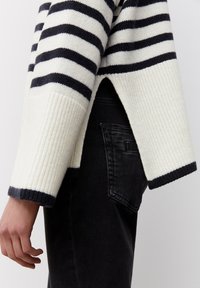 Wzorzysty sweter w kremowo-granatowym kolorze. Ma teksturę ściąganą, szerokie rękawy oraz boczne rozcięcie. Połączony z czarnymi dżinsami.