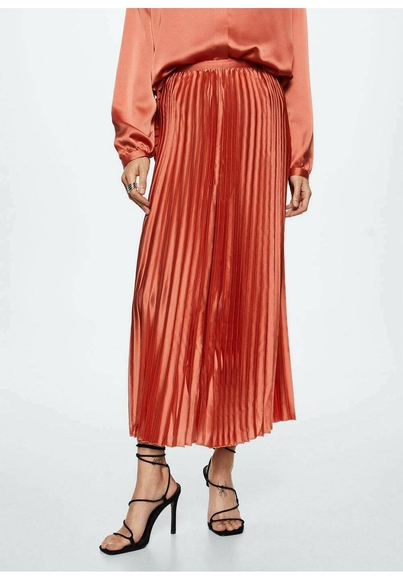 Femme portant une jupe midi plissée brillante couleur rouille avec un chemisier en satin assorti et des sandales noires à talons hauts et à lanières.