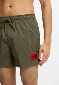 Olivgröna nylonbadshorts med resår i midjan, justerbar dragsko och en röd logotypmärke på vänster lår.