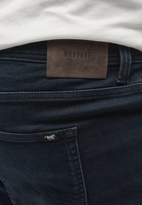 Prim-plan al blugilor denim albastru închis cu o etichetă maro din piele Mustang deasupra buzunarului din spate și un mic logo cu cal pe buzunar.