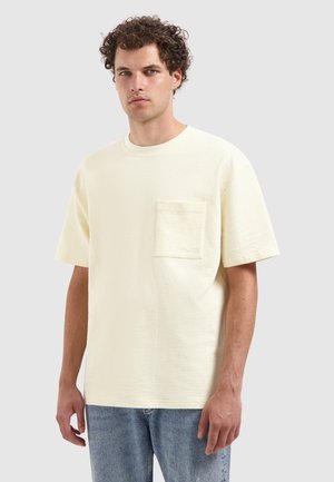 Korte mouwen, ronde hals T-shirt in een zachte, textuurrijke crèmekleurige stof. Voorzien van een enkel borstzakje en een relaxte pasvorm, gecombineerd met lichtblauwe jeans.