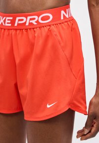 Φωτεινά πορτοκαλί αθλητικά σορτς Nike Pro με ελαστική μέση και λογότυπο Nike σε λευκό χρώμα στο αριστερό πόδι, φορεμένα από άτομο με σκούρο δέρμα.
