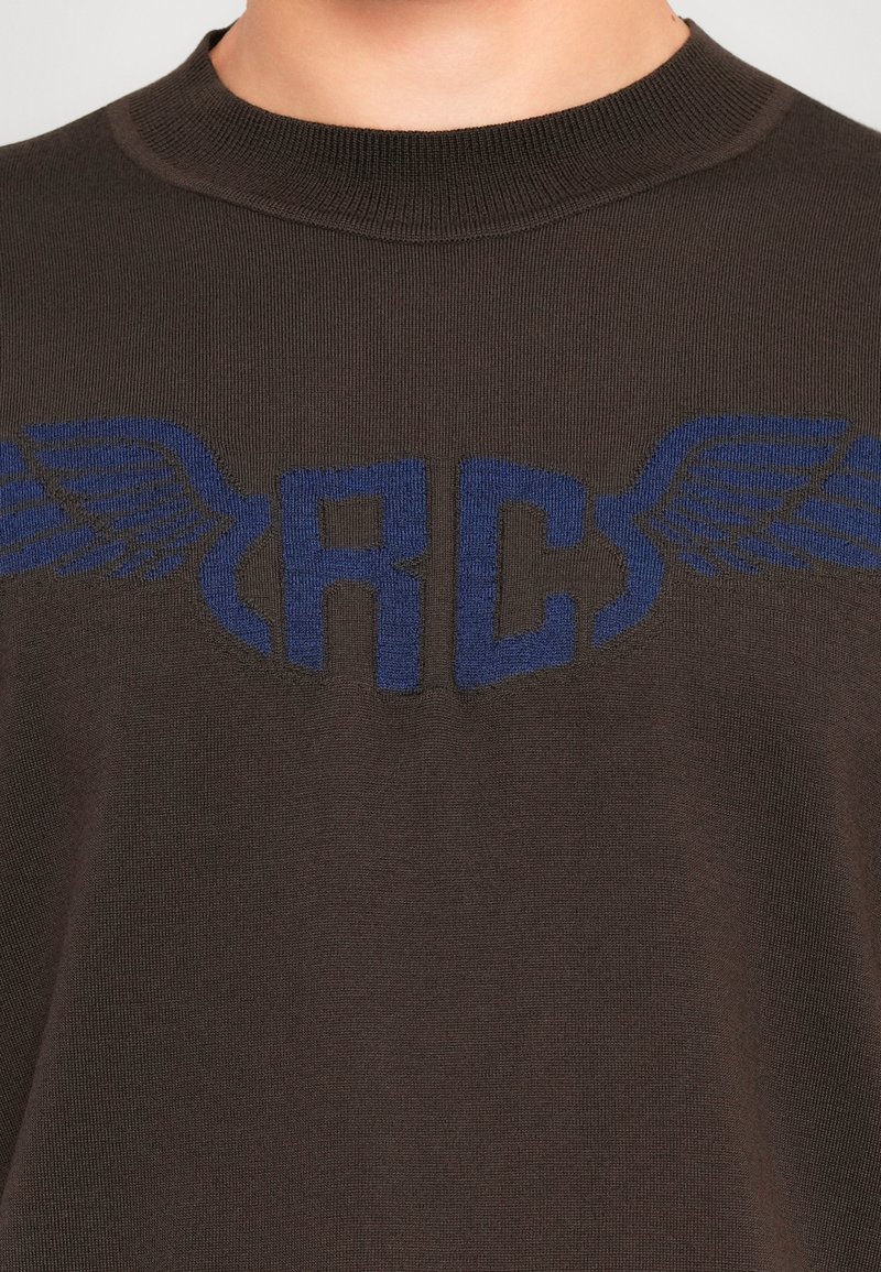 Gros plan sur un pull en laine marron avec des lettres bleues "RC" et des motifs d'ailes sur la poitrine, porté par une personne.