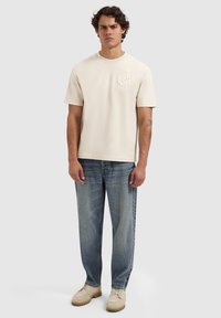 Crème-kleurig katoenen t-shirt met korte mouwen en reliëflogo op de linkerborst, gecombineerd met vervaagde blauwe rechte jeans en beige schoenen.