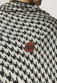 Gehaakte zwart-wit houndsstooth patroon sjaal met een gestructureerde afwerking, voorzien van een ronde bruine logo patch.