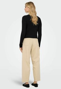 Maglione a coste nero con vita aderente, abbinato a pantaloni ampi beige e scarpe slip-on nere. Sfondo semplice.