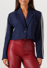 Marineblauwe cropped blazer met één knoop, voorzien van witte gestreepte accenten op de mouwen. Gecombineerd met hooggesneden rode leren broeken.