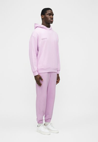Férfi áll, világoslila kapucnis pulóvert és hozzáillő melegítőnadrágot visel, fehér sportcipőben, fehér háttér előtt.