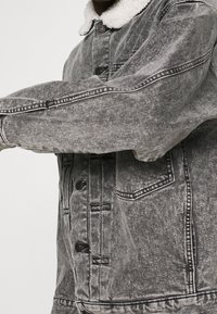 Grå denimjacka med en plyschvit krage, som har en knapplåsning framtill, sidofickor och synliga sömnadsdetaljer.