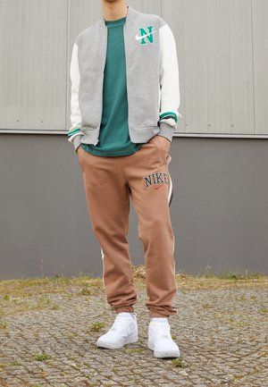 Homme portant une veste varsity grise et blanche, un t-shirt vert, un pantalon de survêtement Nike marron et des baskets blanches, debout sur un sol pavé.