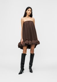 Γυναίκα που φοράει καφέ αμάνικο, φαρδύ φόρεμα με βολάν στο τελείωμα και μαύρες ψηλοτάκουνες μπότες μέχρι το γόνατο, στέκεται μπροστά σε λευκό φόντο.