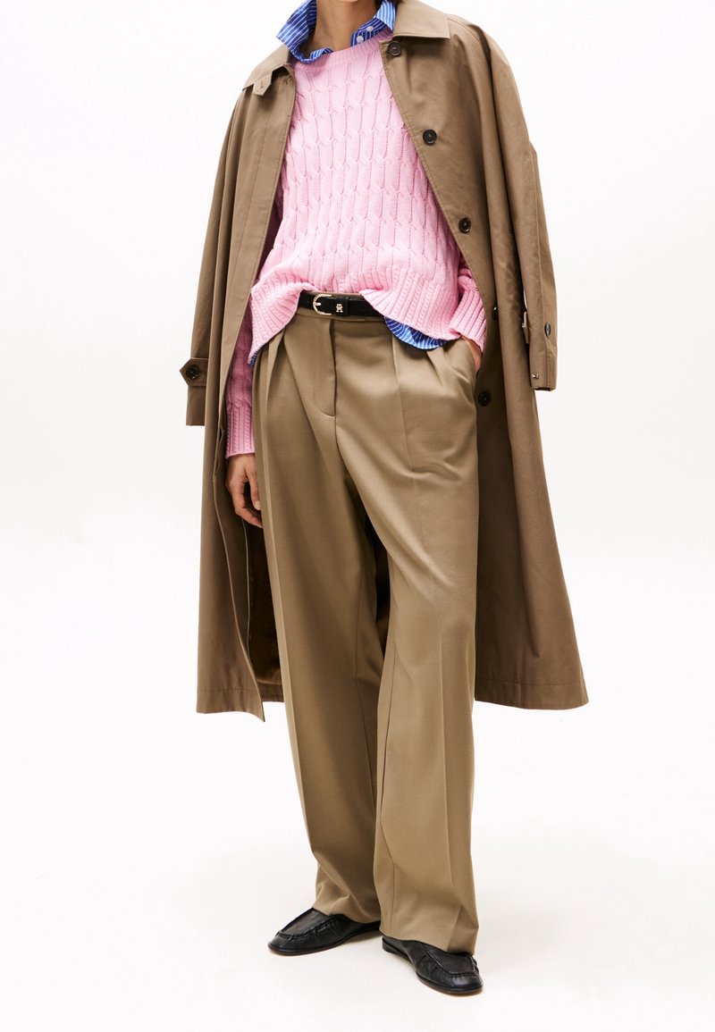 Personne portant un trench-coat beige drapé sur les épaules, un pull en maille torsadée rose par-dessus une chemise rayée bleue, un pantalon plissé beige et des chaussures noires.