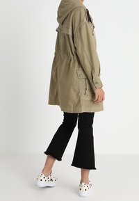 Olive green hooded jacket, mid-thigh length, front pockets, cinched waist. Worn with black flared pants and white shoes with black dots.