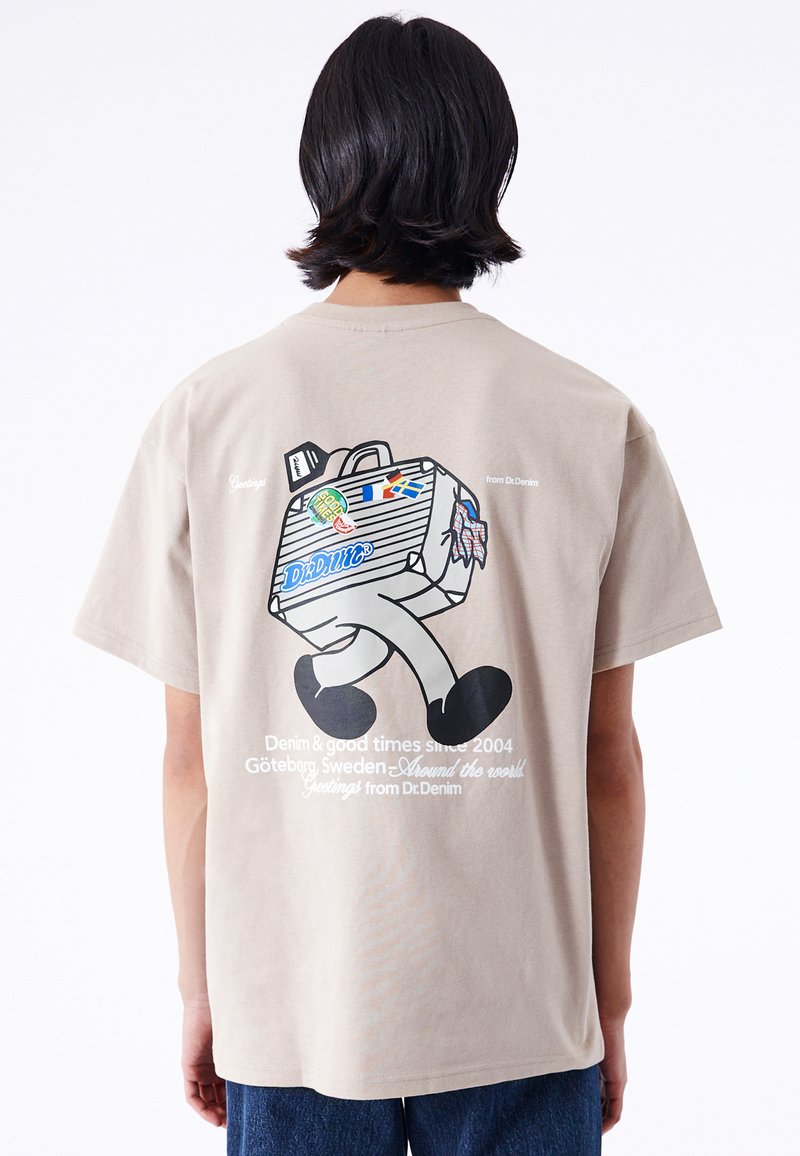 Camiseta de color beige claro con un personaje de dibujos animados caminando con una maleta, el texto "Saludos de Dr.Denim" y varias pegatinas coloridas.