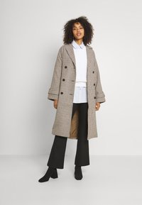 Dvojradový kabát so vzorom houndstooth v hnedej a béžovej farbe, s veľkými gombíkmi, vo voľnom strihu a bočnými vreckami.