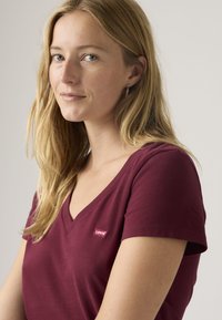 Νεαρή γυναίκα με μακριά ξανθά μαλλιά, φορώντας μπορντό T-shirt Levi's με V λαιμόκοψη, στραμμένη ελαφρώς προς τα αριστερά σε απλό φόντο.