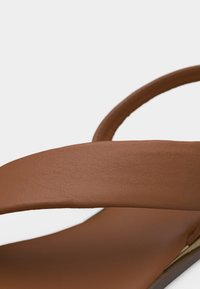 Καφέ δερμάτινο σανδάλι με φαρδιά λουρί πάνω από το πόδι και λουρί στον αστράγαλο, σε απλό φωτεινό φόντο.