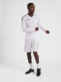 Hvid atletisk langærmet skjorte med sorte chevron-detaljer og matchende shorts, parret med sorte sneakers og hvide sokker.
