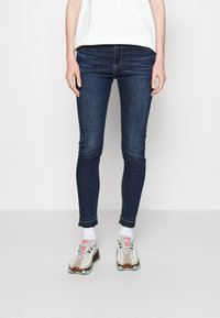 Mörkblå skinny jeans med kontrasterande sömmar, med en midjehöjd och slitna ankelsömmar, tillsammans med beige och grå atletiska sneakers.