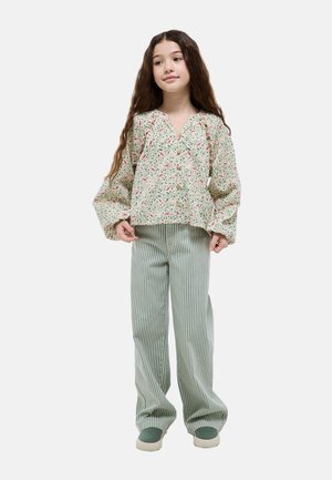Jeune fille aux longs cheveux portant un chemisier boutonné à fleurs, un pantalon large rayé vert et blanc, et des chaussures vertes sans lacets.
