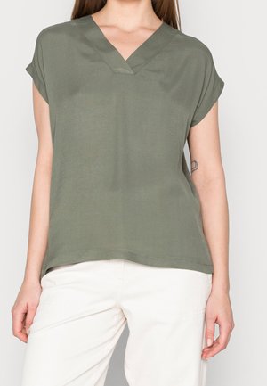 Frau trägt olivgrünes, kurzärmeliges V-Ausschnitt-Oberteil und weiße Hose, zeigt Oberkörper und Mittelteil vor einfachem Hintergrund.